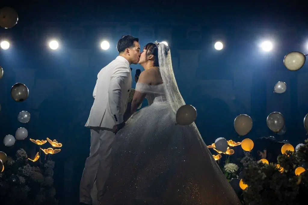 Bride and groom share a romantic kiss on their wedding day surrounded by lights and balloons.