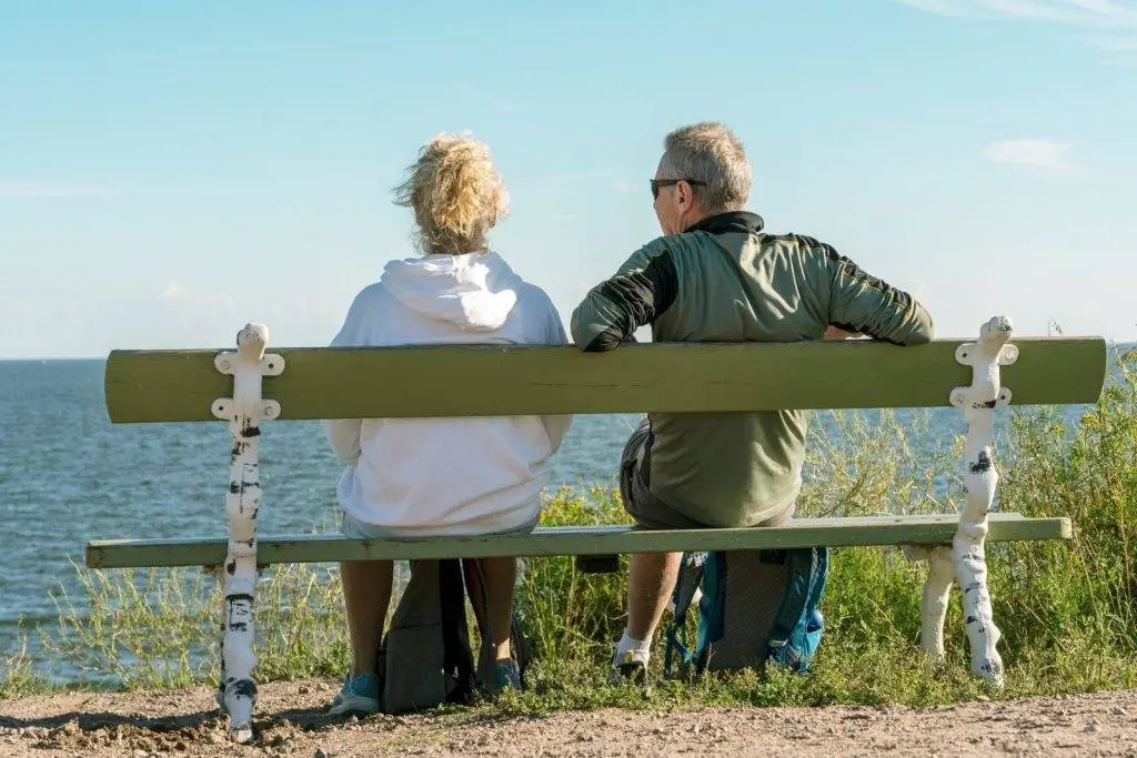 An elderly couple sits on a bench by the sea in Helsinki, enjoying a sunny day.