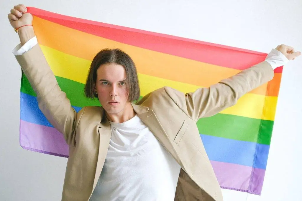 Young man in beige suit proudly holding a rainbow flag symbolizing LGBT pride and identity.