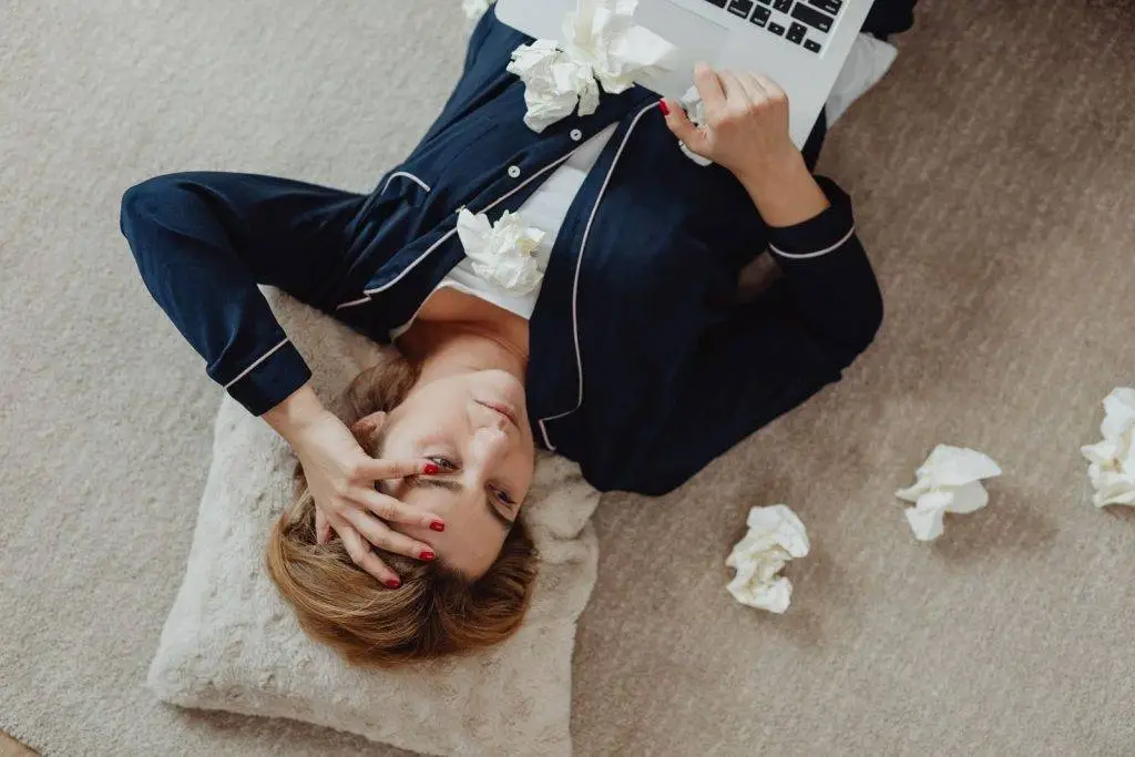 A woman in pajamas, stressed and lying on the floor with a laptop and scattered tissues.