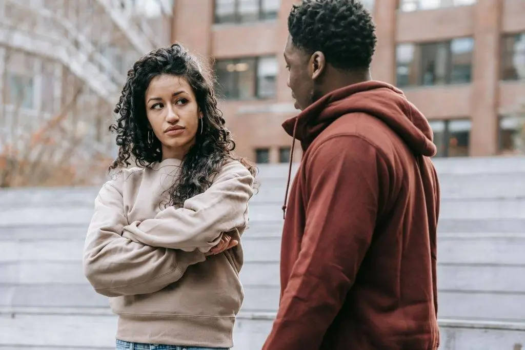 Unset ethnic woman with crossed arms and African American man looking at each other while having quarrel on street with building on blurred background